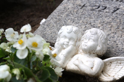 Oberkörper zweier Engel aus weißem Stein lehnen vor einem halb liegendem Grabstein. Vor den Engeln sind weiße Blüten zu sehen. - Copyright: Ev.-Luth. Kirchenkreis Lübeck-Lauenburg